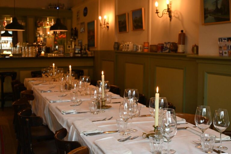 Feestelijk gedekte lange eettafel met brandende kaarsen en glazen in een sfeervol restaurant.