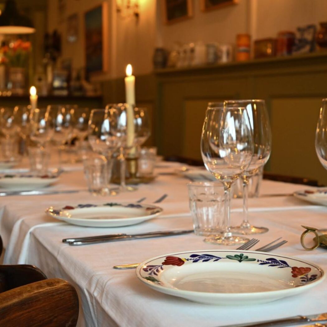 Lange gedekte eettafel in een restaurant met brandende kaarsen, wijnglazen en decoratieve borden.