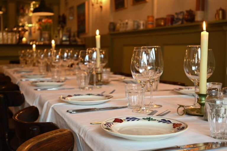 Lange gedekte eettafel in een restaurant met brandende kaarsen, wijnglazen en decoratieve borden.