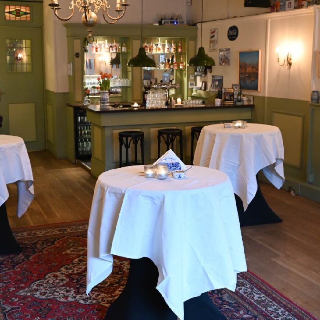 Sfeervolle barruimte met witte sta-tafels, een groene houten bar en rood tapijt op een houten vloer.
