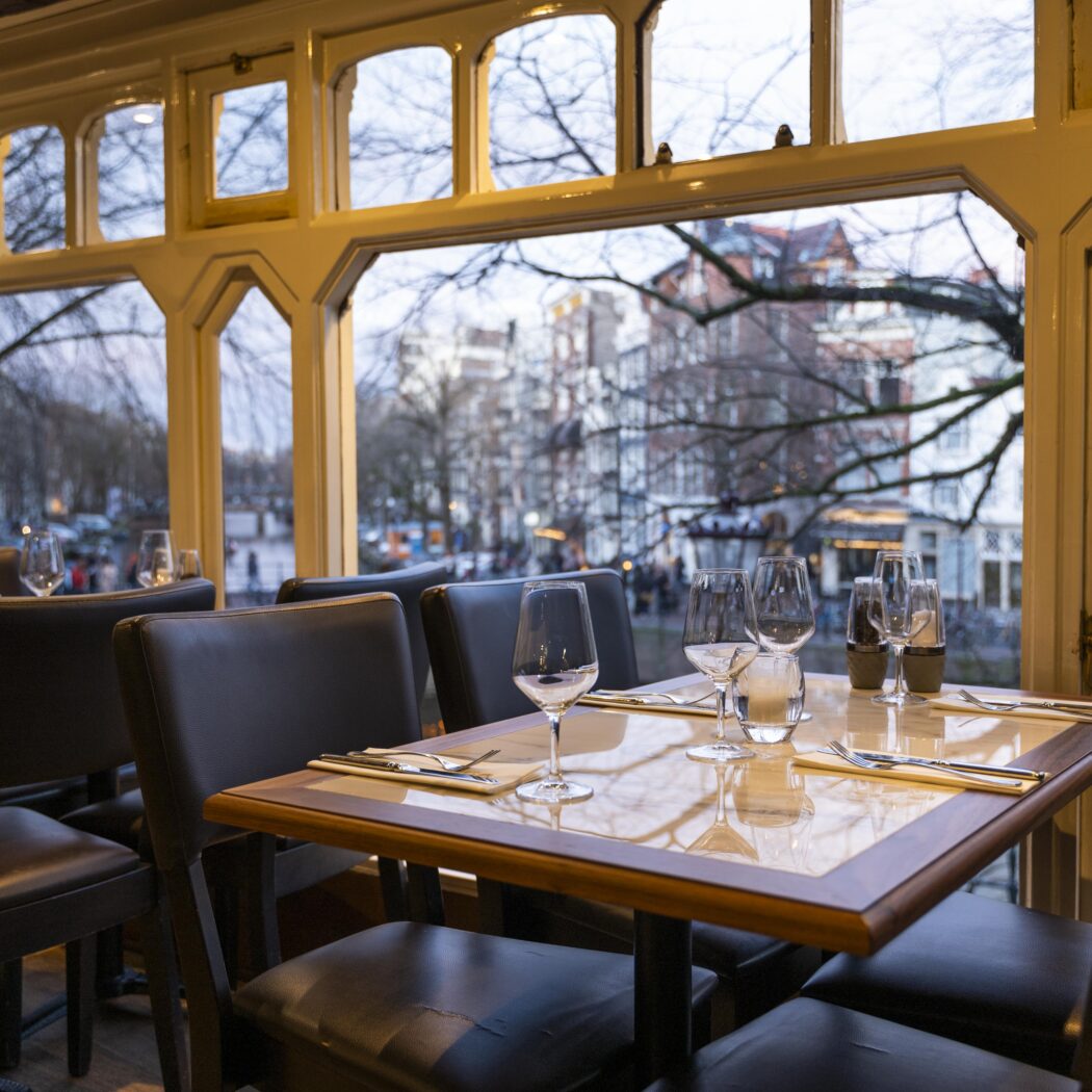 Gedekte tafels en donkere stoelen in een restaurant met grote ramen die uitkijken op een gracht met historische gebouwen.