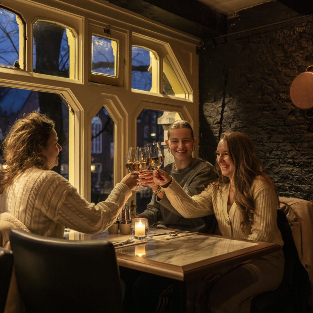 Drie lachende vrouwen proosten met glazen wijn in een sfeervol restaurant, met uitzicht op de stad in de schemering.