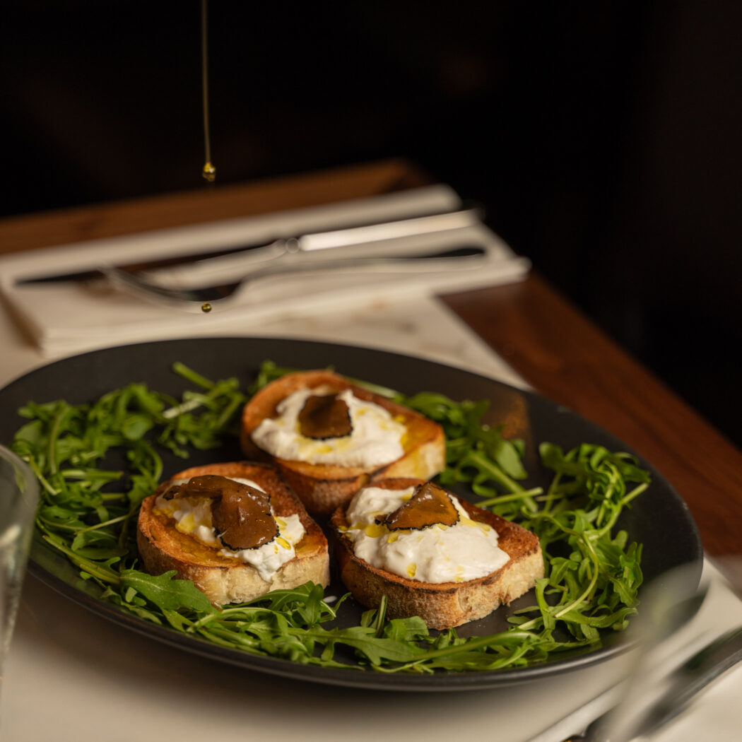 Drie bruschetta met burrata, truffel en rucola op een zwart bord, besprenkeld met olijfolie.