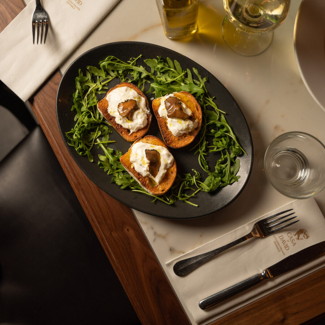 Drie bruschetta met burrata, truffel en rucola op een zwarte schaal, met Casa David servetten op een marmeren tafel.