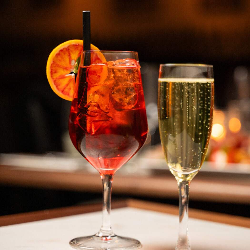 Rode cocktail met bloedsinaasappel en ijs naast een glas fonkelende champagne op een lichte tafel.