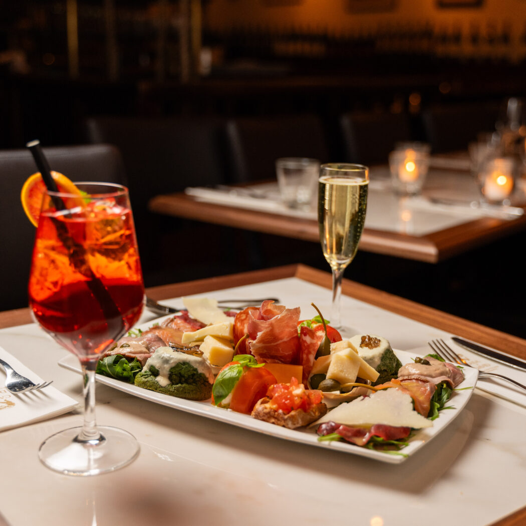 Witte schotel met antipasti, rode cocktail en champagneglas op een lichte tafel in een sfeervol restaurant.