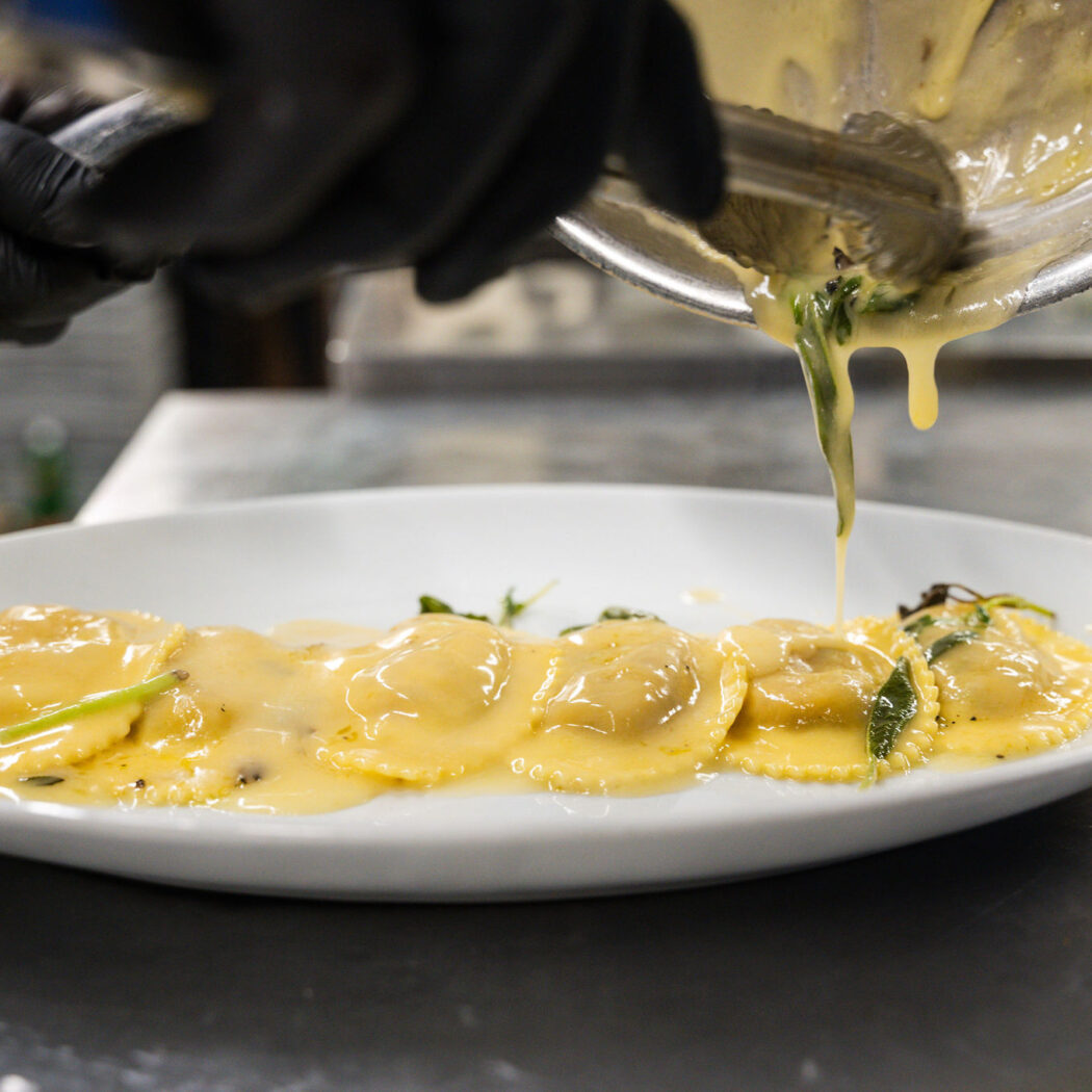 Een hand met zwarte handschoen giet romige saus over ravioli met kruiden op een wit bord.