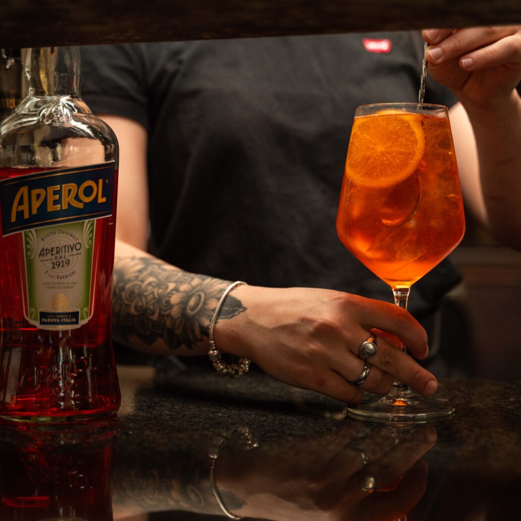 Een getatoeëerde hand roert in een Aperol Spritz met sinaasappel en ijs op een donkere bar, naast een fles Aperol Aperitivo 1919.