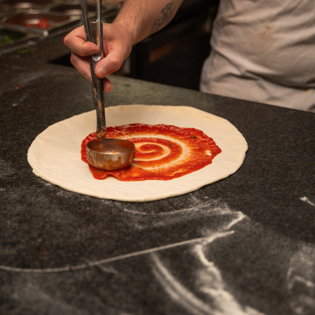 Een kok smeert met een lepel rode pizzasaus in een spiraal op vers pizzadeeg.