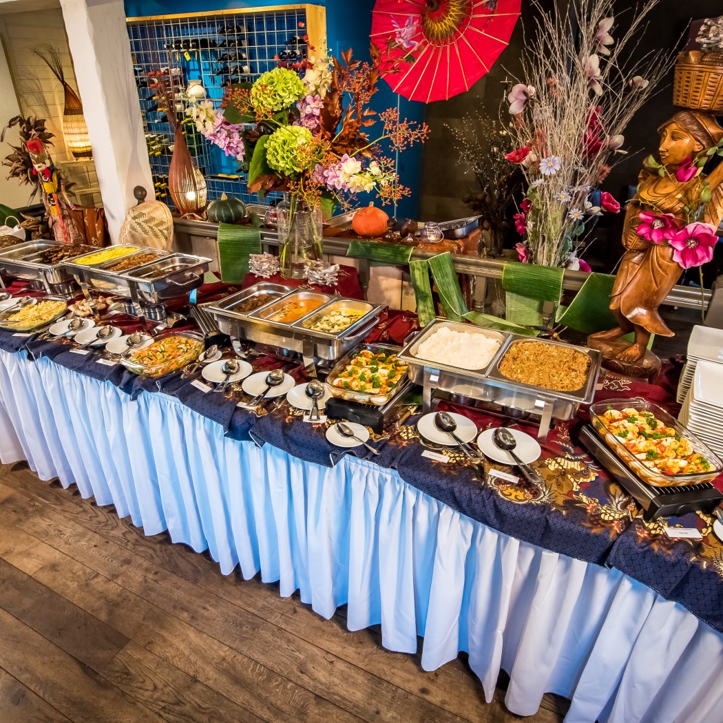 Uitgebreid buffet met warme gerechten op een tafel met batik-kleed, omgeven door Aziatische decoraties en een houten beeld.
