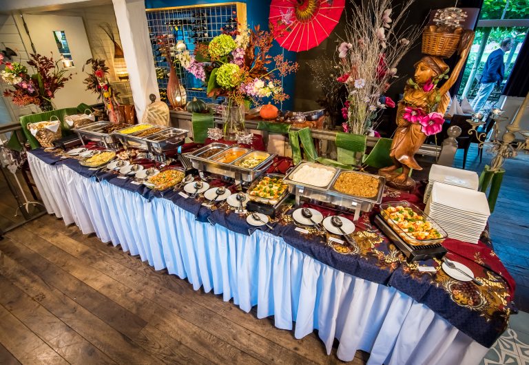 Uitgebreid buffet met warme gerechten op een tafel met batik-kleed, omgeven door Aziatische decoraties en een houten beeld.