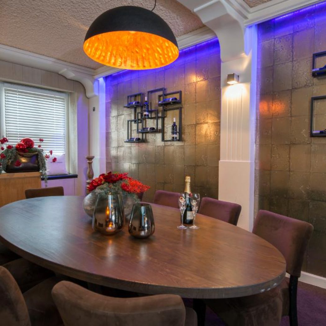 Elegante eetruimte met ovale houten tafel, paarse stoelen, bronzen tegelwand met blauwe ledverlichting en designlamp.
