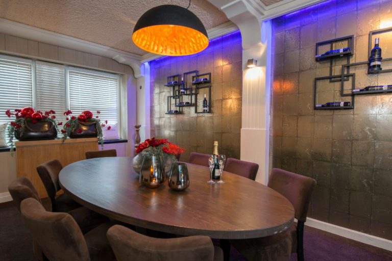 Elegante eetruimte met ovale houten tafel, paarse stoelen, bronzen tegelwand met blauwe ledverlichting en designlamp.