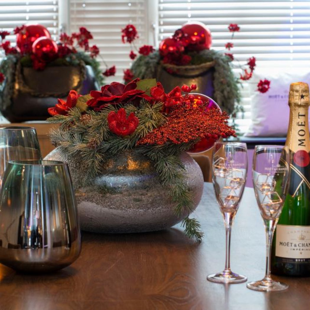 Moët & Chandon champagnefles, twee flutes, rood feestelijk bloemstuk en zilveren vazen op een houten tafel.