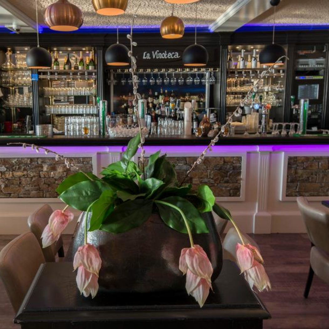 Sfeervolle bar van La Vinoteca met paarse LED-verlichting, flessen, glazen en een plant met hangende roze bloemen.