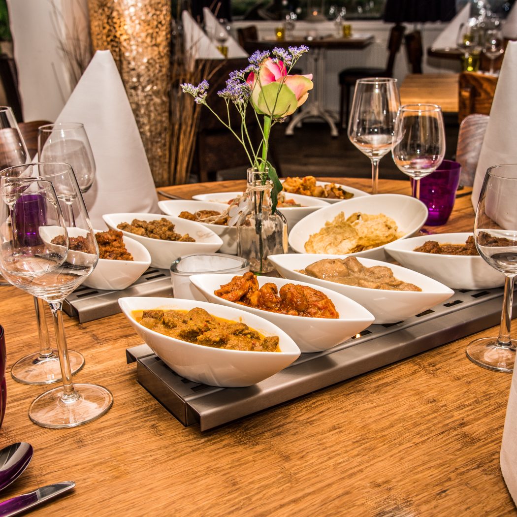 Houten restauranttafel met een schaal vol Indische gerechten, wijnglazen, paarse glazen en bloemen.