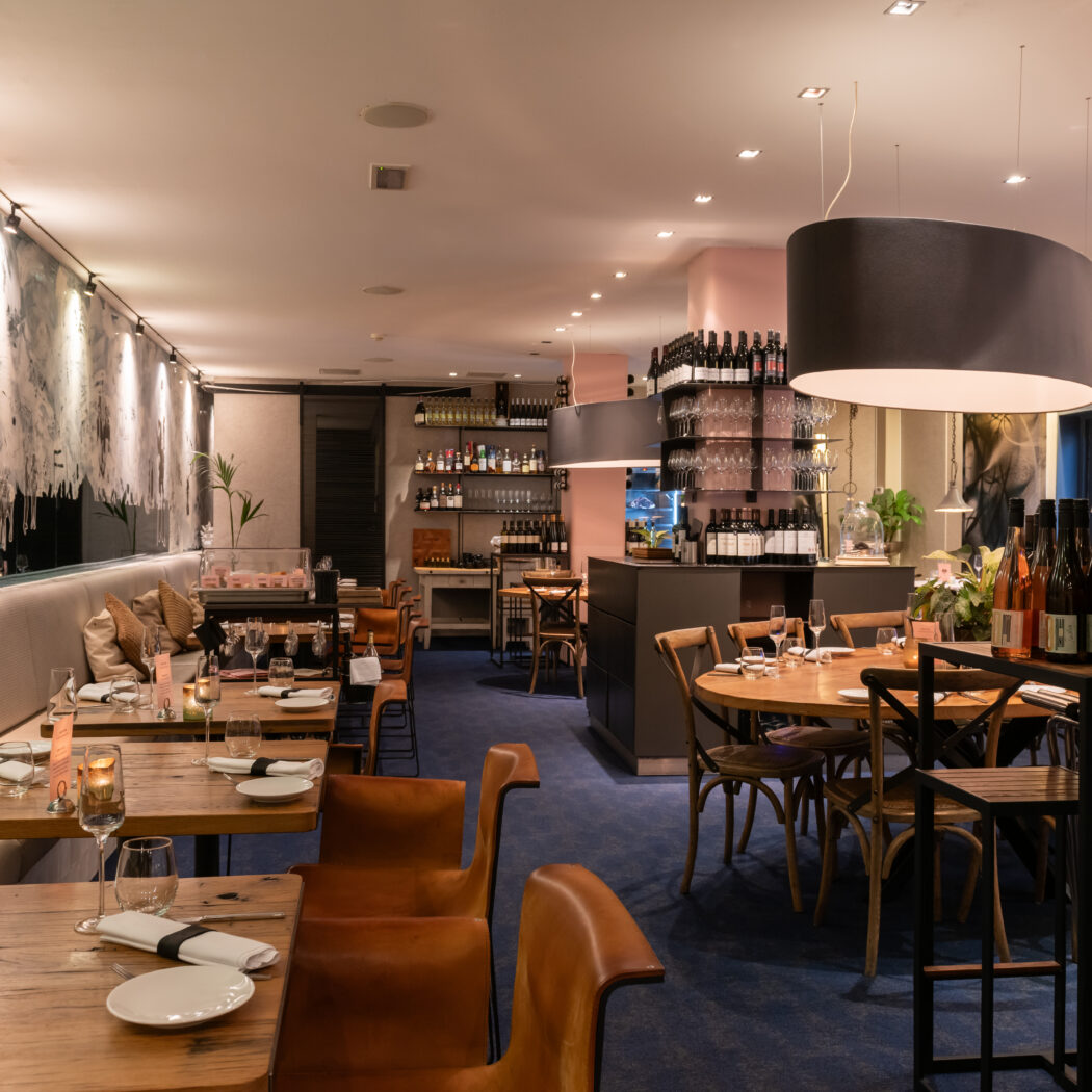 Sfeervol restaurantinterieur met opgedekte houten tafels, roze wanden en flessen wijn op rekken en een kar.