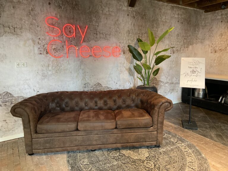 Bruine Chesterfield bank voor een ruwe muur met rood Say Cheese neonlicht, een plant en Take a Pic bord.