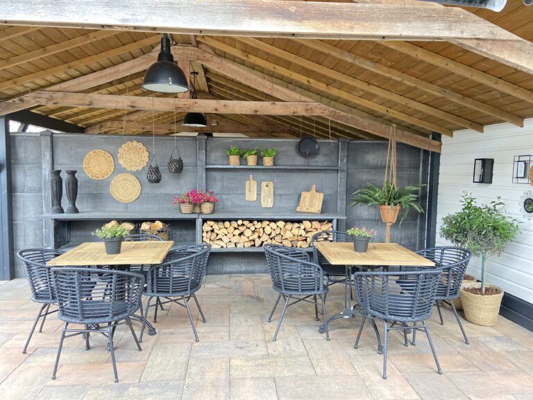 Gezellige buitenruimte onder houten overkapping met twee tafels, donkere stoelen, planten en gestapeld haardhout.