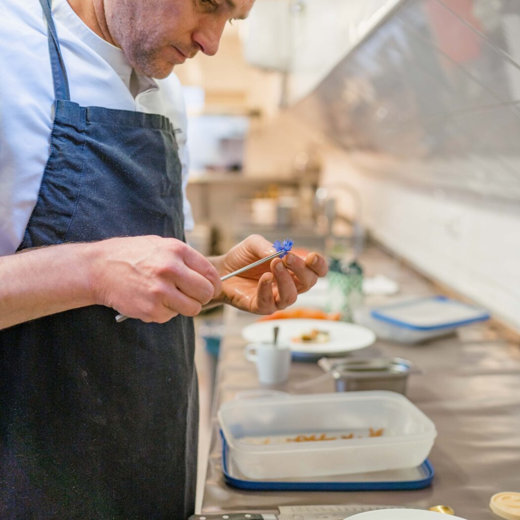 Chef in zwarte schort decoreert een dessert met een klein blauw bloemetje en een pincet.