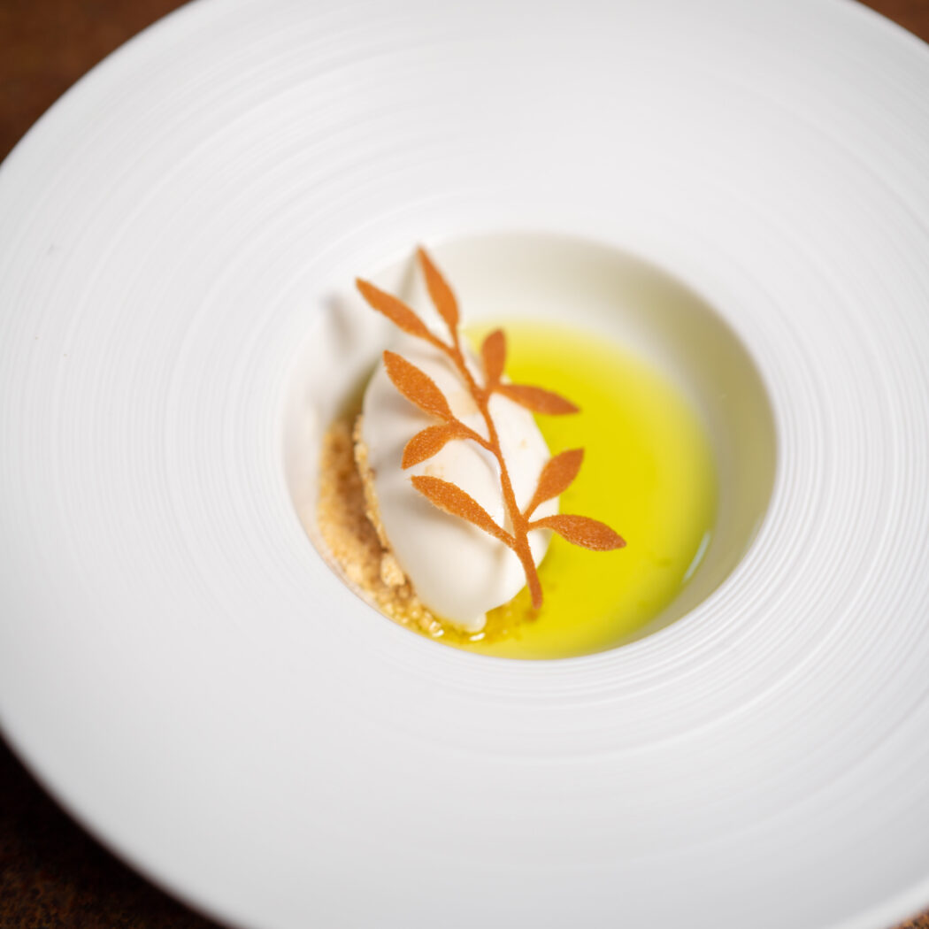 Culinair dessert in witte kom met witte mousse, kruimelbodem, lichtgele vloeistof en een oranje blad decoratie.