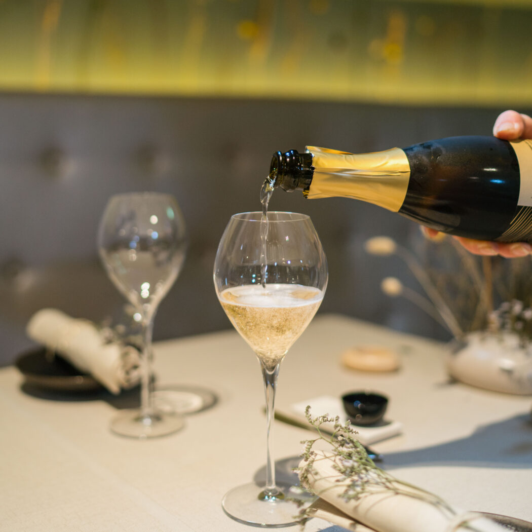 Een hand schenkt sprankelende wijn uit een fles met gouden folie in een glas op een gedekte tafel.
