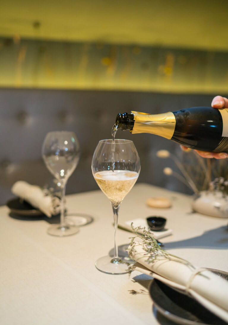Een hand schenkt sprankelende wijn uit een fles met gouden folie in een glas op een gedekte tafel.