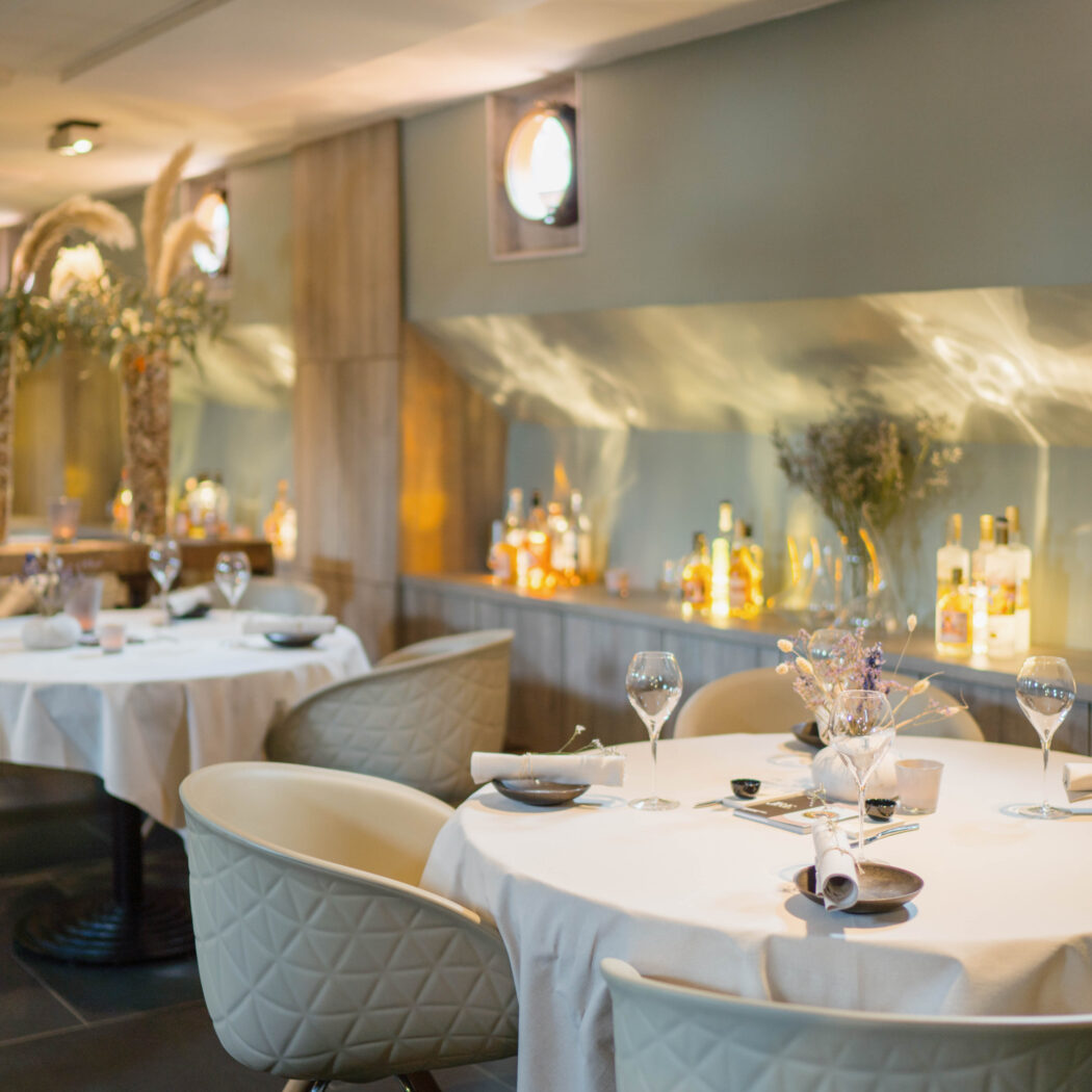 Elegant restaurantinterieur met gedekte ronde tafels, beige stoelen en een sfeervolle bar.