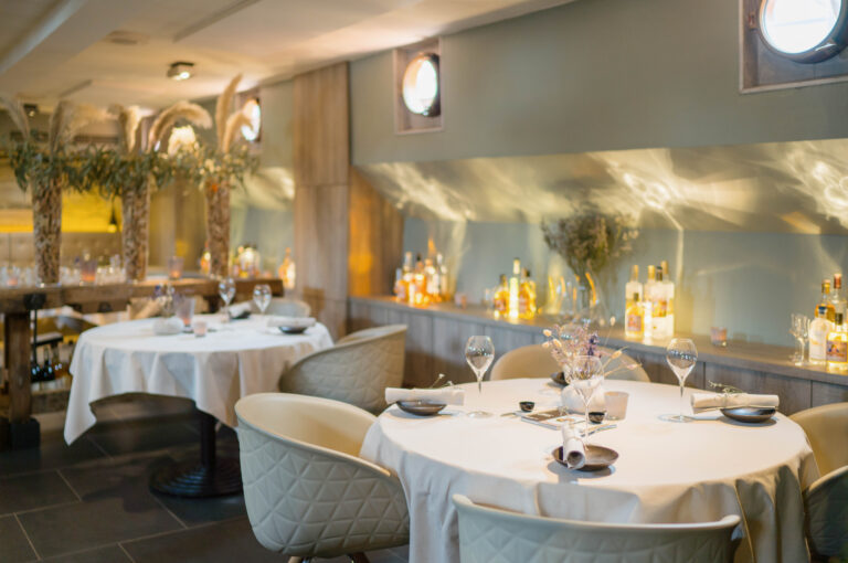 Elegant restaurantinterieur met gedekte ronde tafels, beige stoelen en een sfeervolle bar.