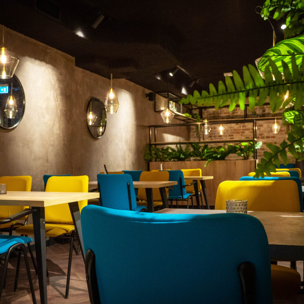 Modern restaurant interieur met blauwe en gele stoelen, houten tafels, planten en warm licht.