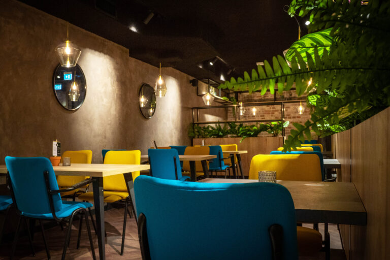 Modern restaurant interieur met blauwe en gele stoelen, houten tafels, planten en warm licht.