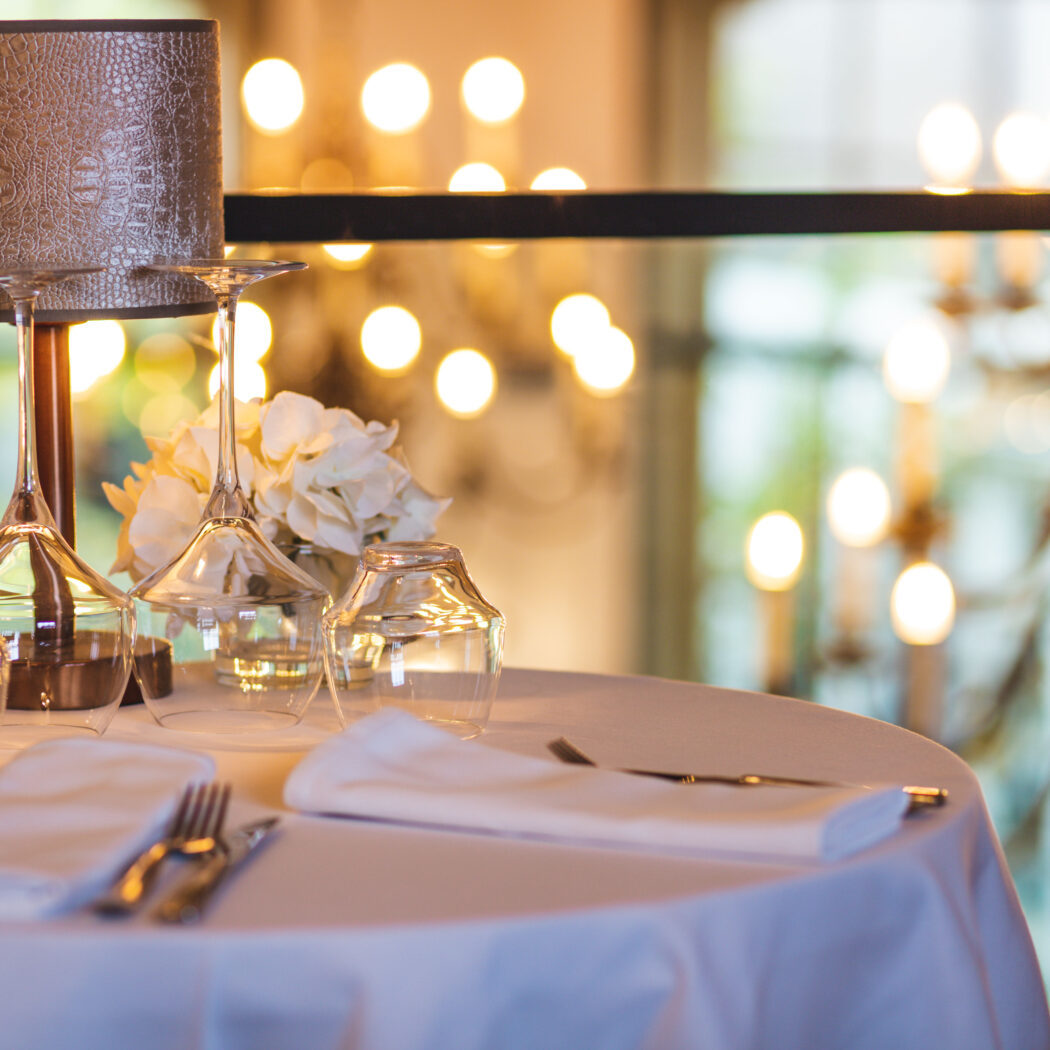 Gedekte tafel met wit linnen, glazen, bestek, tafellamp en witte bloemen in een sfeervol restaurant.