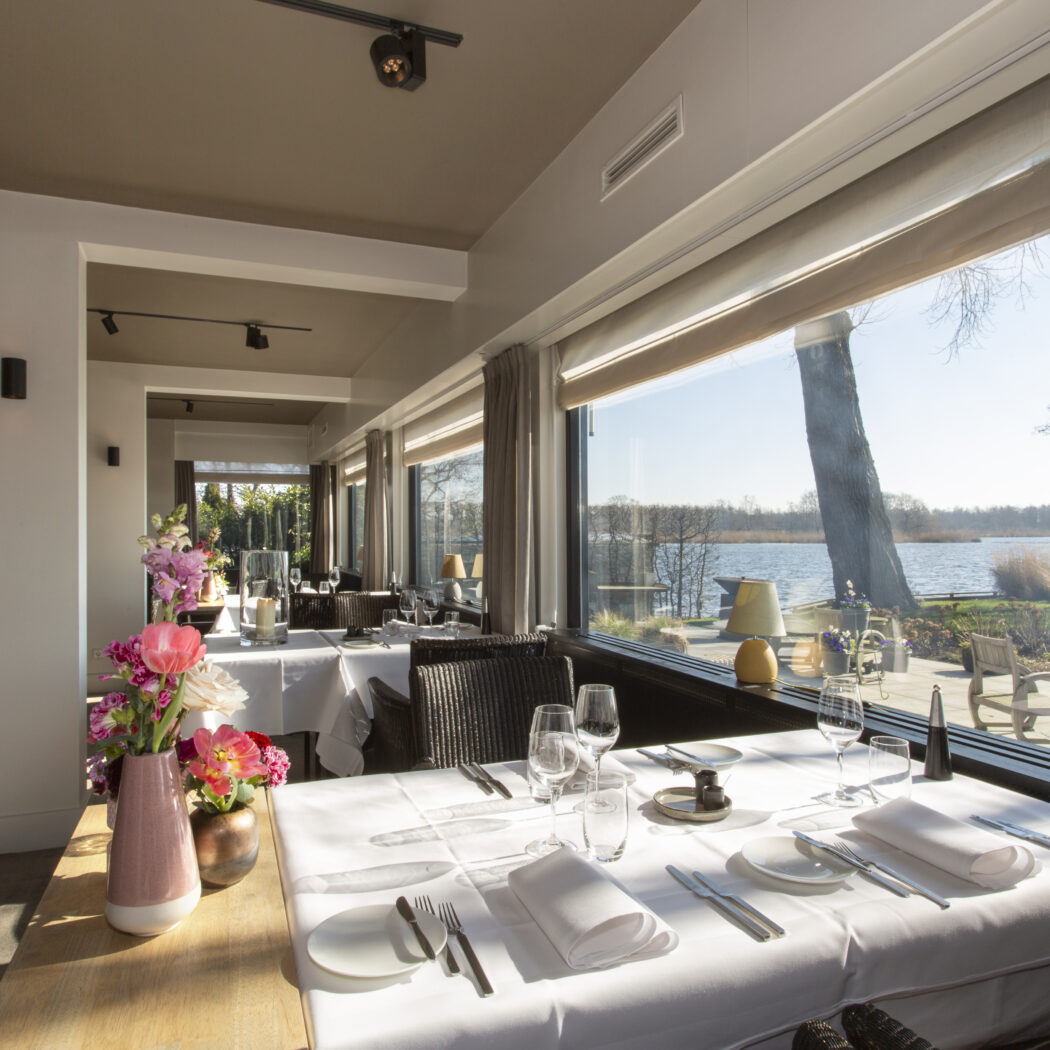 Elegante restaurantzaal met wit gedekte tafels, bloemdecoratie en prachtig uitzicht over het water.
