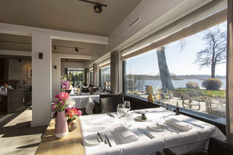 Elegante restaurantzaal met wit gedekte tafels, bloemdecoratie en prachtig uitzicht over het water.