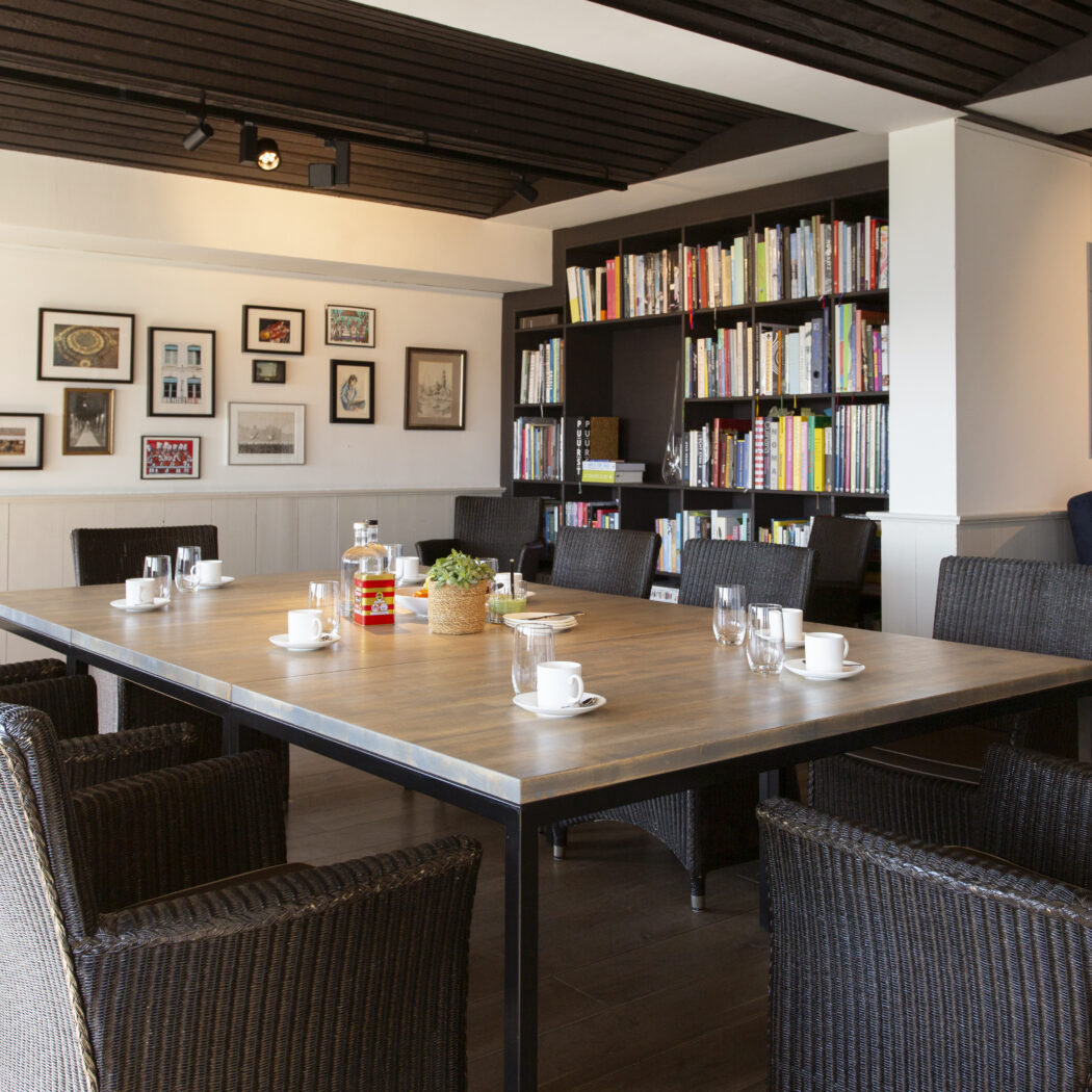 Lange houten tafel met donkere gevlochten stoelen in een kamer met een grote boekenkast en galerijwand.