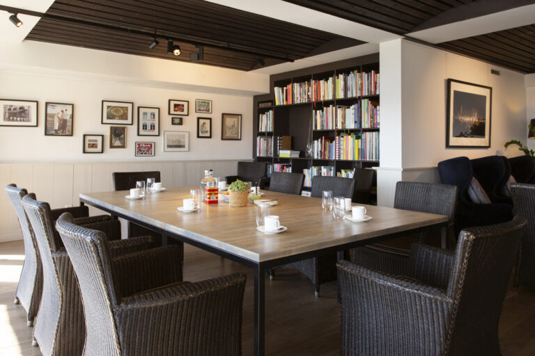 Lange houten tafel met donkere gevlochten stoelen in een kamer met een grote boekenkast en galerijwand.