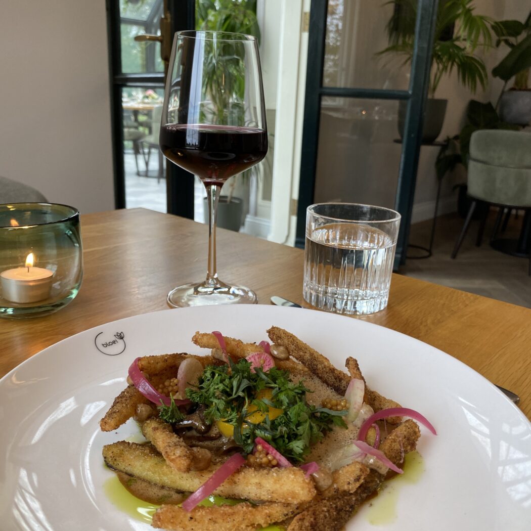 Krokant gerecht met rode ui, groene kruiden en ‘bloei’ logo, met rode wijn en water op een houten tafel in een restaurant.