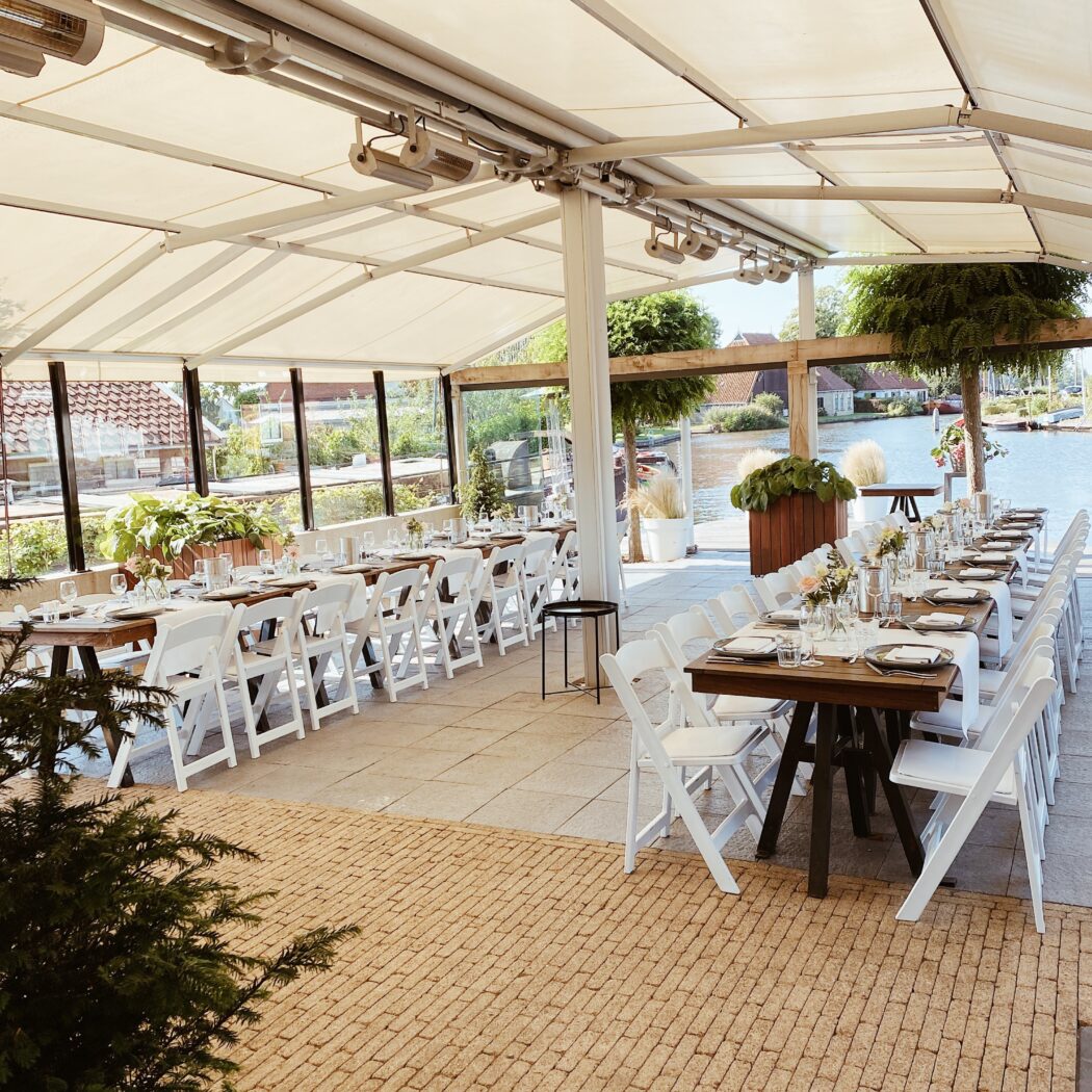 Lange gedekte houten tafels met witte stoelen op een ruim terras aan het water, klaar voor een evenement.