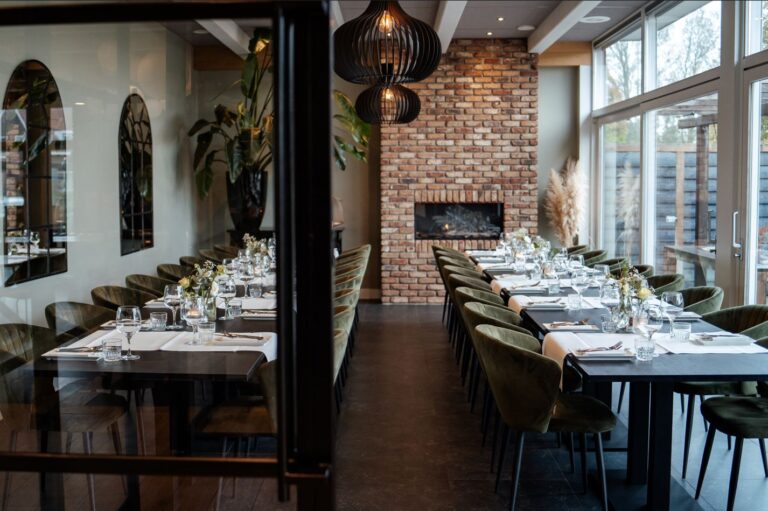 Een lange, elegant gedekte eetzaal met donkergroene fluwelen stoelen, sfeervolle verlichting en een bakstenen haard.