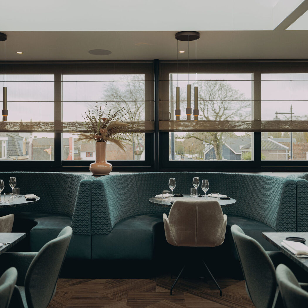 Stijlvol restaurant met groenblauwe boothbanken, gedekte donkergrijze tafels en hoge ramen.