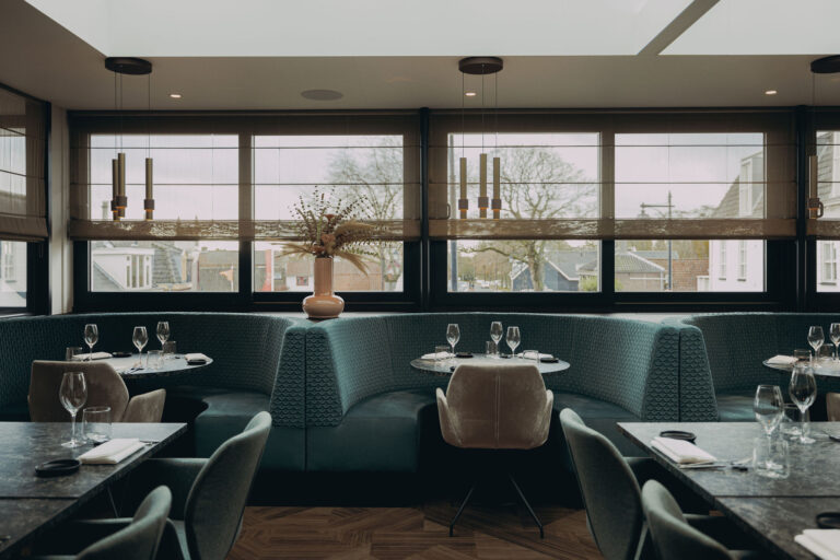 Stijlvol restaurant met groenblauwe boothbanken, gedekte donkergrijze tafels en hoge ramen.
