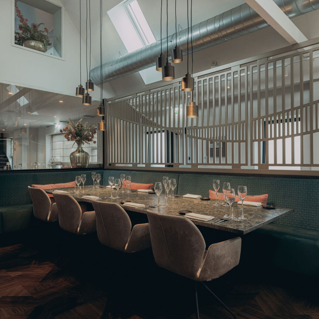 Sfeervol restaurant interieur met lange eettafel, groene bank, beige stoelen en moderne hanglampen.