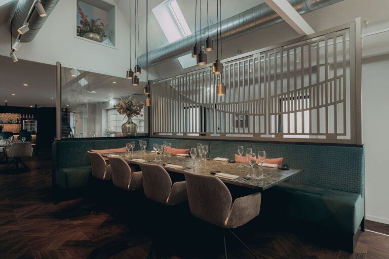 Sfeervol restaurant interieur met lange eettafel, groene bank, beige stoelen en moderne hanglampen.