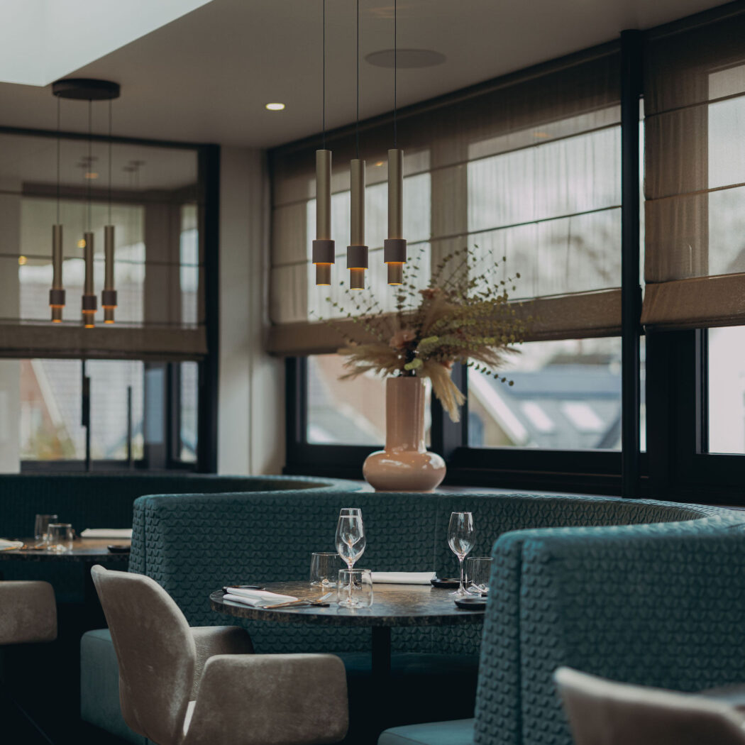 Modern restaurant interieur met gebogen teal banken, opgedekte tafels, gouden hanglampen en vaas met droogbloemen.