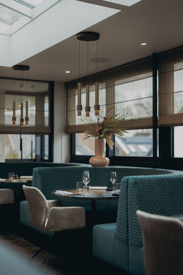 Modern restaurant interieur met gebogen teal banken, opgedekte tafels, gouden hanglampen en vaas met droogbloemen.