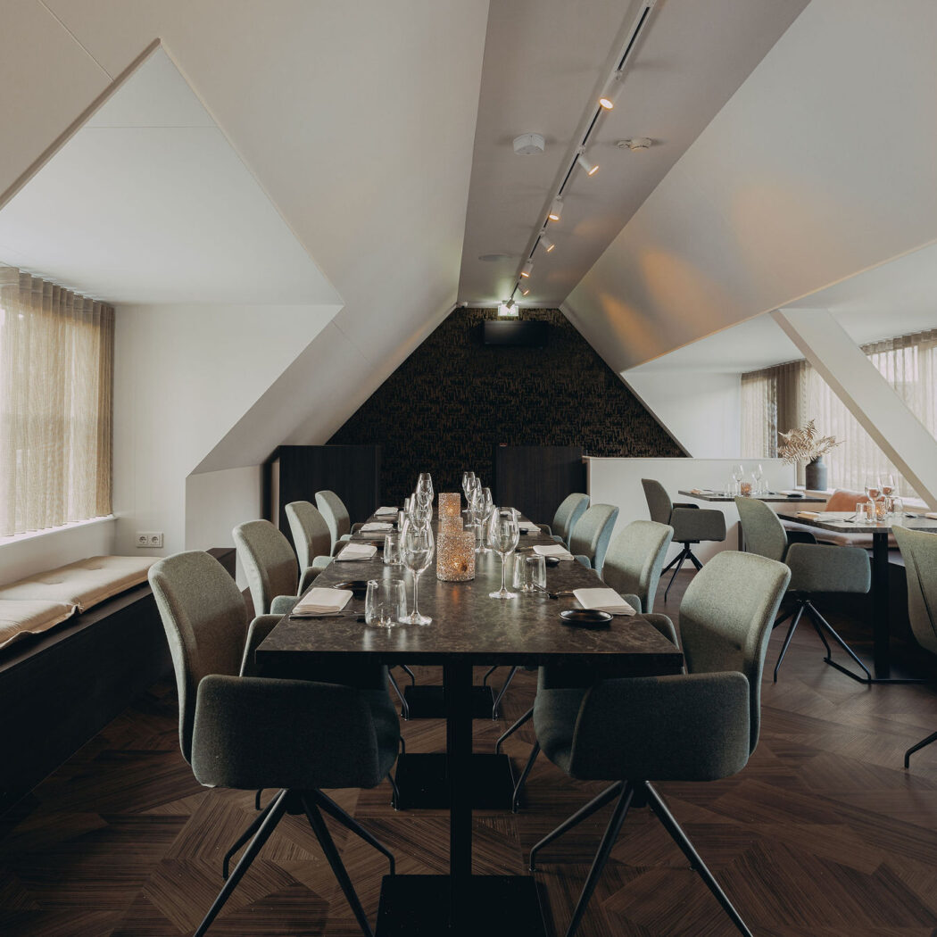 Stijlvol restaurantinterieur met lange gedekte tafel, comfortabele stoelen en een raam met zitbank.