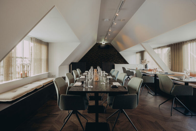 Stijlvol restaurantinterieur met lange gedekte tafel, comfortabele stoelen en een raam met zitbank.