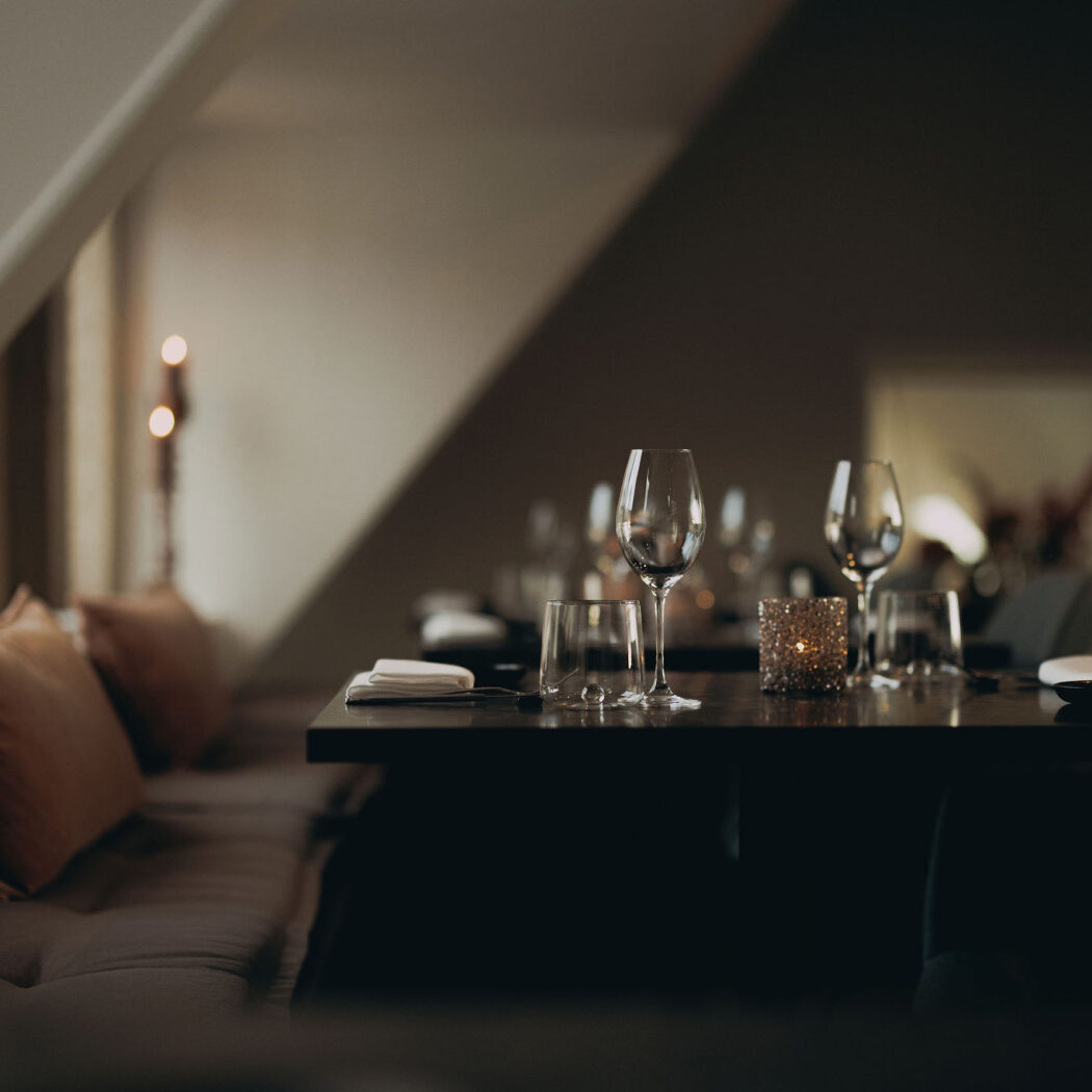 Sfeervolle gedekte tafel met wijnglazen, kaarslicht en zachte kussens in een schemerig restaurant.