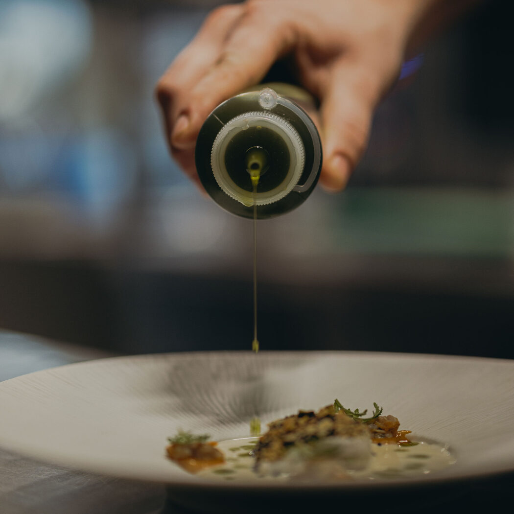 Hand van een chef die olijfolie uit een groene fles over een elegant gerecht druppelt.