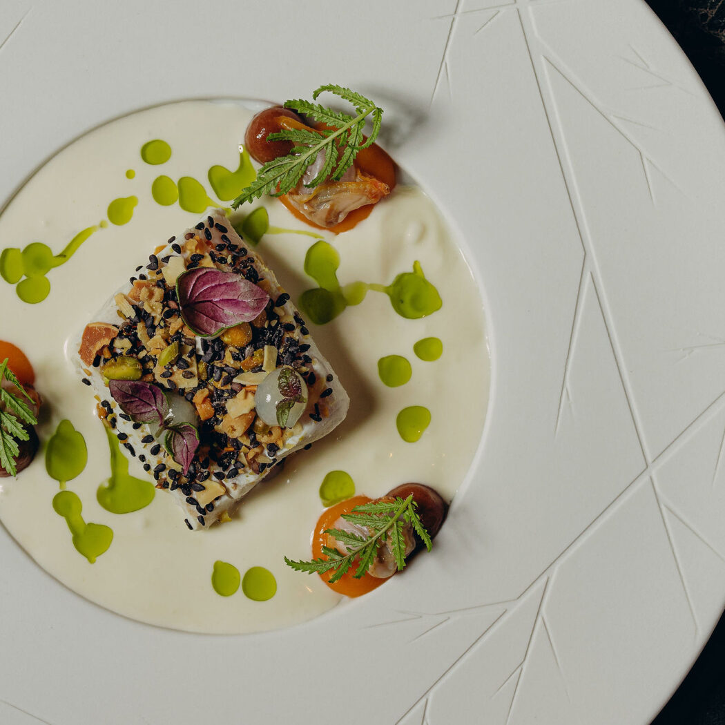 Fijnproevers visgerecht met sesam, noten, oranje puree en groene olie op wit bord met bladreliëf.